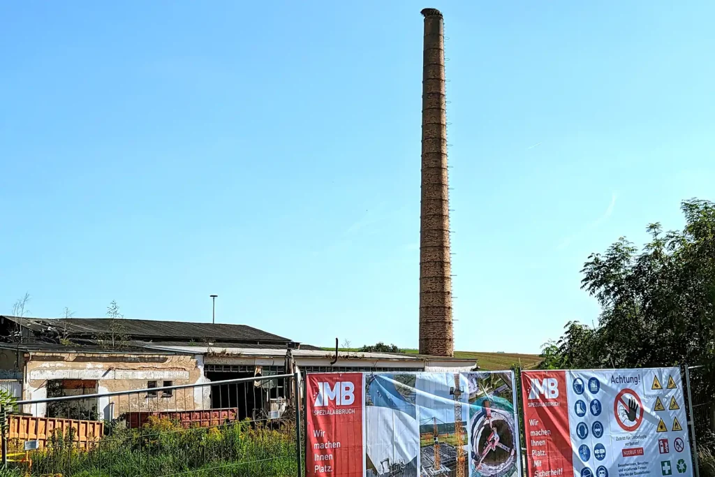 Blick vom Bauzaun mit Planen von MB-Spezialabbruch auf den hohen Ziegelschornstein